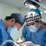 Two cardiac surgeons wearing sterile scrubs, surgical masks, and caps performing an intricate open heart surgery procedure under bright operating room lamps