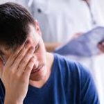 Stressed man holding his head in his hand while a healthcare professional holds a medical clipboard in the background