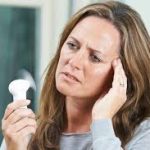 Mid-adult woman experiencing a menopause hot flash holding a small portable fan to her face while touching her temple in discomfort