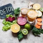 A collection of fresh fruits and vegetables, including leafy greens, beets, citrus, and glasses of fresh juice, arranged on a surface with a "Detox" sign.