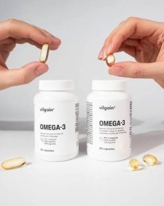 Hands holding golden Omega-3 softgel capsules above two white bottles of Omega-3 dietary supplements.